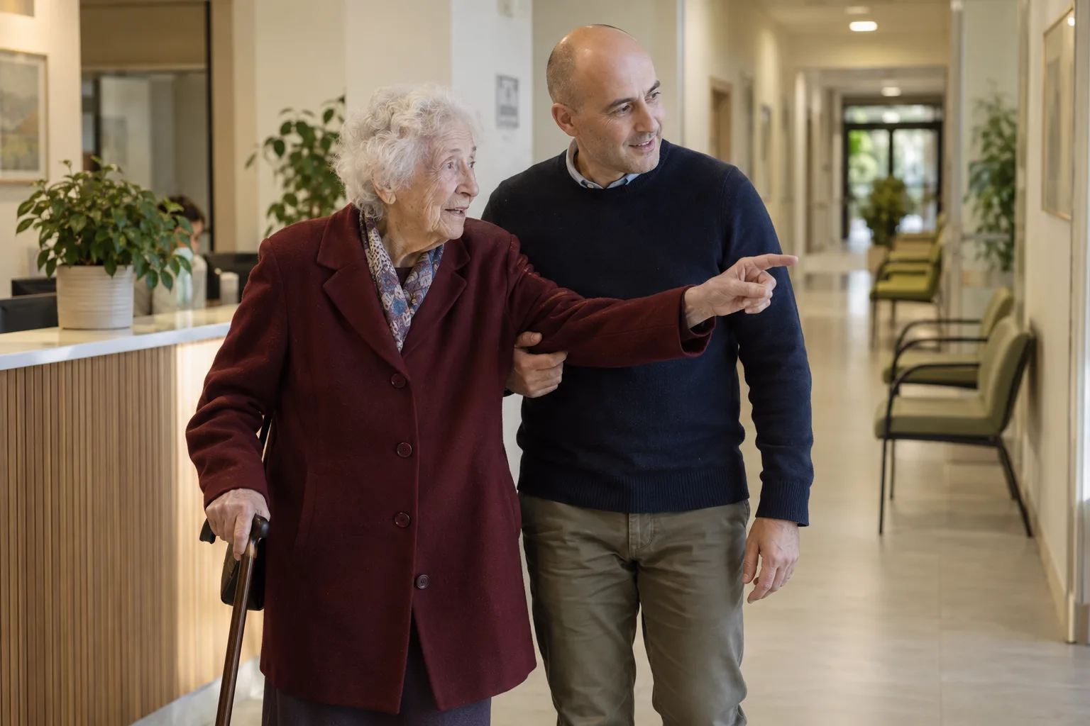 Una donna anziana con il bastone viene accompagnata lungo il corridoio di un ambulatorio.