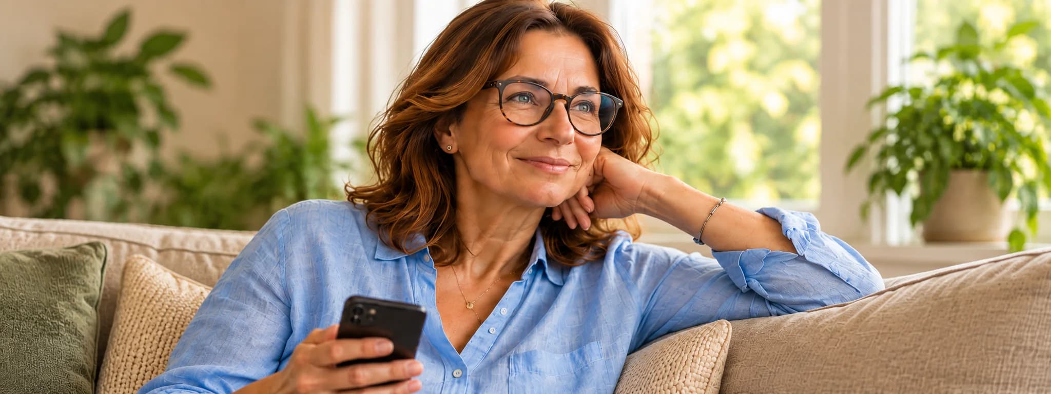 Una donna seduta sul divano di casa, con il telefono in mano, che guarda fuori dalla finestra e sorride.